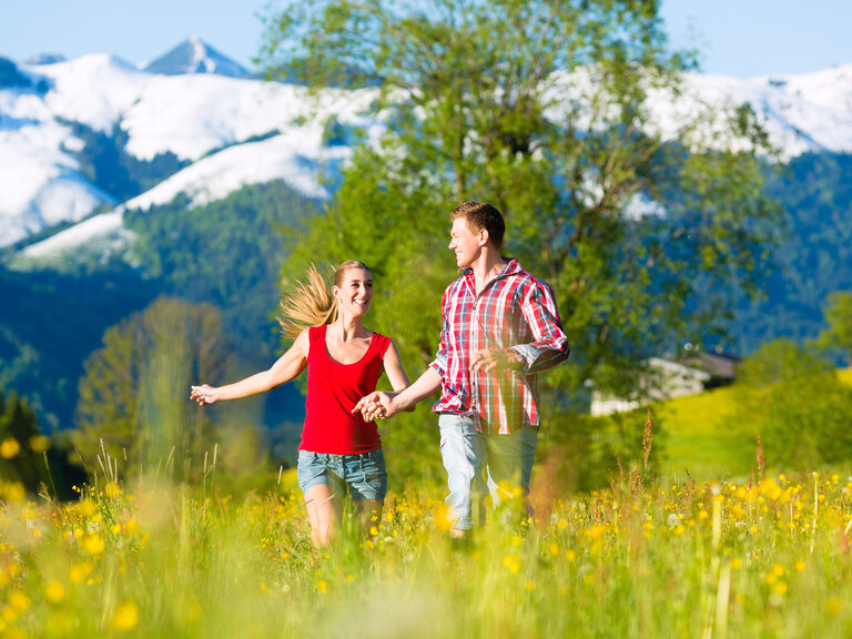 Ein Paar läuft lachend über eine Wiese während im Hintergrund die schneebedeckten Berge glitzern.